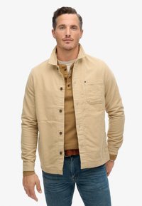 Beige katoenen jack met een kraag, knopen aan de voorkant en een borstzak. Draagt over een lichtbruine henley-shirt en blauwe jeans.