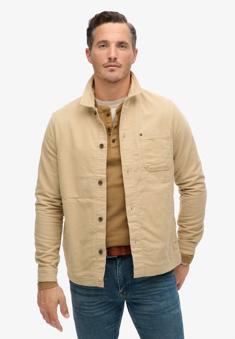 Beige katoenen jack met een kraag, knopen aan de voorkant en een borstzak. Draagt over een lichtbruine henley-shirt en blauwe jeans.