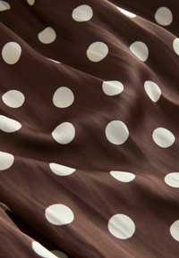 Tessuto marrone con una superficie liscia, caratterizzato da grandi pois bianchi disposti in un motivo regolare. Il tessuto si drappeggia dolcemente, creando pieghe delicate.