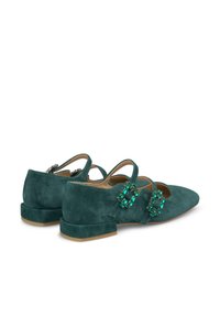 Zapatos planos de ante en color azul verdoso con dos tiras que presentan acentos de piedras preciosas verdes, punta afilada y tacón cuadrado bajo. Textura suave, diseño elegante.