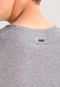 Gros plan sur le haut du dos d'une personne portant un pull en tricot gris avec une petite étiquette métallique de marque près de l'encolure.