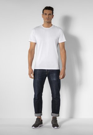 Hvid T-shirt med rund halsudskæring, blå denim jeans med opkrøllede ben, og grå sneakers med lilla detaljer, sat mod en neutral baggrund.