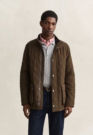 Jeune homme portant une veste matelassée marron par-dessus un pull gris à torsades et une chemise rose à col, debout devant un fond clair uni.