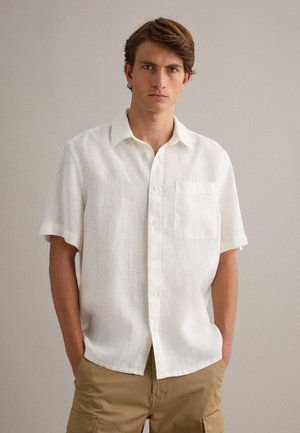 Jeune homme aux cheveux bruns courts portant une chemise blanche à manches courtes à boutons et un short cargo beige, debout les mains dans les poches.