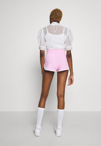 Bílá průsvitná blůza s volnými rukávy a klíčovou dírkou na zádech, v kombinaci s růžovými šortkami s bílým lemem. Model nosí bílé tenisky a ponožky.