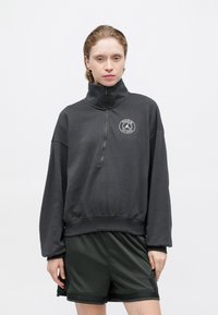 Donkergrijze zip-up sweatshirt met hoge kraag, ribbelmanchetten en een circulair Parijs-logo op de borst. Draagt een combinatie met donkergroene shorts.