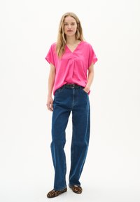Blusa rosa a scollo a v, con maniche corte, abbinata a jeans a vita alta di colore blu scuro. La modella indossa scarpe con stampa leopardo. Sfondo pulito e minimale.