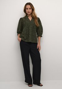 Grøn bluse med blonde detaljer, V-hals og pufærmer, parret med sorte brede bukser. Blusen har blomster mønstre og struktureret stof.