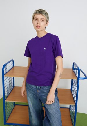 Camiseta de algodón morada con cuello redondo y pequeño logo blanco en el pecho, combinada con pantalones vaqueros de pierna ancha azul claros. Diseño sencillo.
