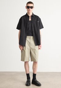 Camisa negra de manga corta con botones sobre una camiseta negra de tirantes, combinada con pantalones cortos beige y zapatos negros, con calcetines negros de canalé.
