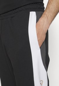 Pantalones de chándal negros con una franja blanca en el lateral y detalles en gris. Elaborados con una tela suave, con cinturilla elástica y bolsillo lateral oculto.