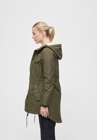 Olive green parka with a hood, featuring a soft inner lining, side pockets, and adjustable drawstrings at the hem. Vertical zipper detail visible.