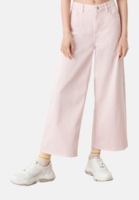 Jean large rose clair en denim, doté d'une fermeture à boutons et de poches avant, porté avec des baskets blanches et des chaussettes rayées.