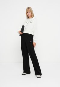 Weiße zip-up Kapuzenjacke, kombiniert mit schwarzen weit geschnittenen Hosen mit einem strukturierten Muster und gesticktem Logo. Schwarze Tasche, graue Schuhe.