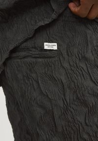 Crni strukturirani materijal s naboranim uzorkom. Na odjeći je vidljiva bijela oznaka s natpisom "JACK&JONES EST.1990".