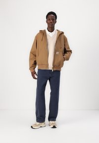 Veste à capuche marron sur un pull crème, associée à un jean bleu foncé et des baskets beiges. Le tissu semble texturé et décontracté.