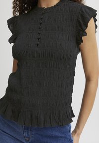 Haut noir texturé avec un design smocké, des manches volantes, un col haut et six boutons décoratifs sur le devant. Ourlet légèrement évasé.