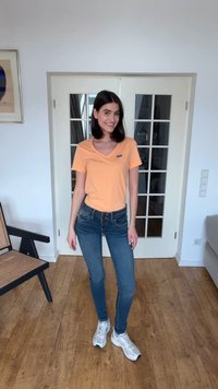 Noor naine, tumedate juustega, kannab oranži t-särki, siniseid teksaseid ja valgeid tenniseid, seisab puidust põrandal valgete topeltustega uste ees.