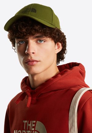 Grüne Baseballkappe mit gebogenem Schirm, mit einem Logo auf der Vorderseite, kombiniert mit einem roten Hoodie, der eine Kordel und einen Schulterriemen zeigt.