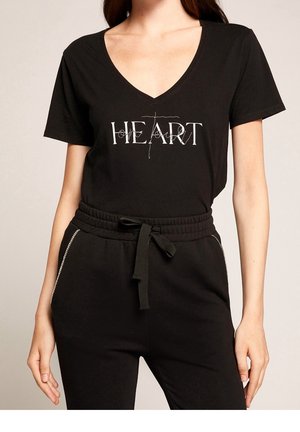 Vrouw draagt een zwart V-hals T-shirt met de tekst "HEART" en een kruisontwerp, gecombineerd met zwarte broek met trekkoord en strassdetails.