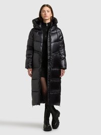 Lange zwarte donsjas met een glanzende afwerking, ritssluiting aan de voorkant, grote capuchon en zijzakken. Draagt over een zwarte gebreide jurk en stevige laarzen.