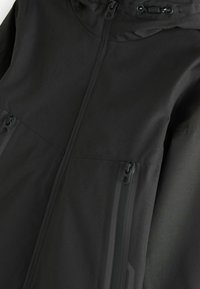 Sort vandtæt jakke med frontlynlås, lynlåslommer i siden og justerbar hætte med snore.