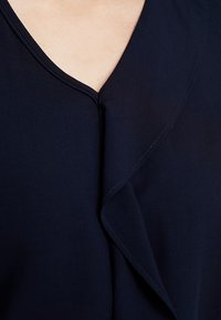 Marinblus med V-ringning, som har en mjuk, flödig fall och subtila sömdetaljer längs kanterna. Slät tygtextur.