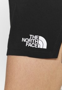 Detailní záběr na stehno osoby, která má na sobě černé šortky s bílým logem "The North Face" vytištěným v blízkosti lemu.