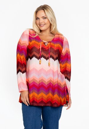 Kleurvolle langemouwen tuniek met een zigzagpatroon in tinten roze, oranje en rood. Heeft een veterhalslijn met kraalaccenten.