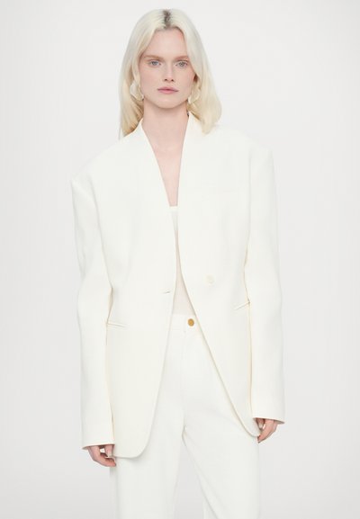 Blazer blanc surdimensionné avec un col châle, fermeture à un bouton et poches avant, associé à un haut et un pantalon de couleur claire.