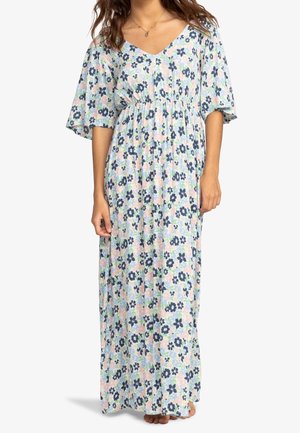 Femme portant une longue robe fleurie ample avec des manches courtes larges dans des tons pastel, debout pieds nus sur un fond uni.