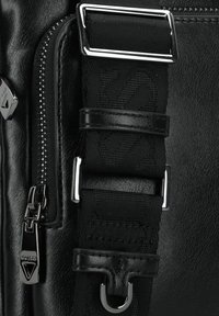 Dettaglio di una borsa in pelle nera con cerniera, accenti metallici e una tracolla testurizzata con fibbia regolabile. Il tessuto è liscio con un morbido sheen.