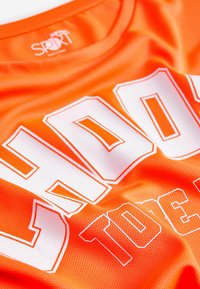 Lys orange sportsjersey med meshtekstur, med stor hvid skrift. Crew neck design med kontrasterende accenter. Letvægtsmateriale.