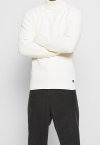 Cremig gerippter Strickrollkragenpullover mit langen Ärmeln, der ein figurbetontes Design aufweist und ein kleines Markenetikett am Saum hat. Kombiniert mit dunklen Cordhosen.