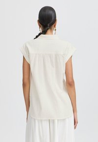 Camicetta corta beige chiaro con maniche corte, colletto, righe verticali e orlo curvo con dettagli arricciati sul retro.