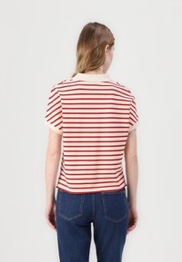 Pruhované tričko v krémové barvě se vodorovnými červenými pruhy, krátkými rukávy a pevným límečkem. Vyrobeno z měkké, lehké tkaniny.