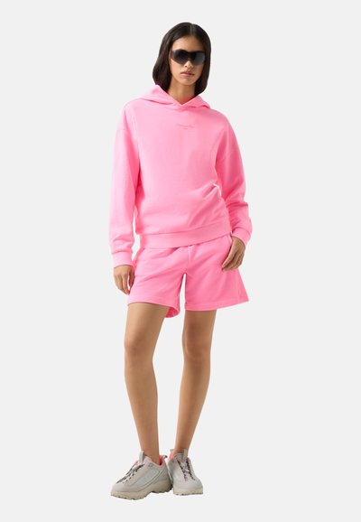 Femme portant un sweat à capuche rose vif et un short assorti, de grandes lunettes de soleil noires et des baskets blanches, debout devant un fond uni.
