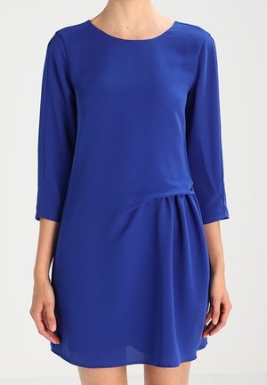 Femme portant une robe bleu roi longueur genou avec des manches trois-quarts et un détail drapé sur le côté droit.