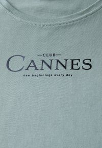 Světle zelené bavlněné tričko s kulatým výstřihem, na kterém je černý potisk s nápisem "CLUB CANNES" a frází "nové začátky každý den."
