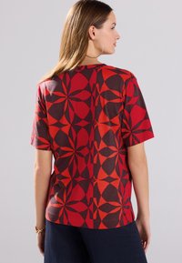 Short-sleeve red shirt with geometric patterns in shades of red and dark gray. Soft fabric with a relaxed fit, featuring a round neckline.