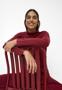 Burgunderfarbener Rollkragenpullover mit glatter Textur und Strassverzierungen an den Ärmeln, getragen von einer Person, die auf einem passenden roten Stuhl sitzt.