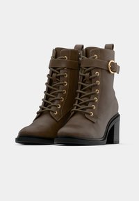 Καφέ δερμάτινα μποτάκια αστραγάλου με χρυσές λεπτομέρειες, κορδόνια μπροστά, φερμουάρ στο πλάι και χοντρό τακούνι. Λείος υφής με ανθεκτική κατασκευή.