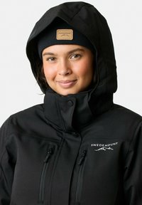 Zwarte softshell jas met een rits aan de voorkant, twee borstzakken en een logopatch. De capuchon is verstelbaar en gevoerd met een warme stof.