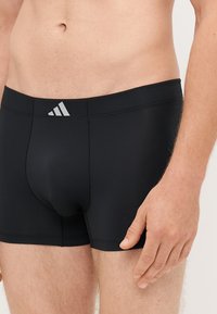 Calções de banho pretos com um ajuste apertado, feitos de material elástico. Apresentam um logo branco da Adidas na cintura e detalhes de design sem costuras.