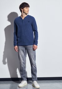 Jongeman staat tegen een witte muur, draagt een blauw langetermijn henley-shirt, vervaagde grijze spijkerbroek en witte sneakers, en kijkt opzij.