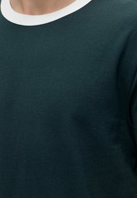 Κοντινό πλάνο ενός ατόμου που φορά πράσινο σκοτεινό μπλουζάκι με λευκή γιακά, δείχνοντας το πάνω μέρος του στήθους και τον λαιμό.
