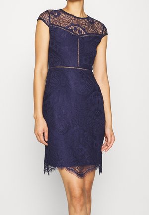 Mujer vestida con un vestido azul marino de encaje hasta la rodilla, con mangas cortas y detalles de encaje transparente en el escote y el dobladillo.