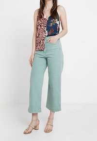 Flerfärgad topp med leopardmönster och blommönster, kombinerad med ljusgröna vida byxor. Sandalerna har en blockklackdesign.