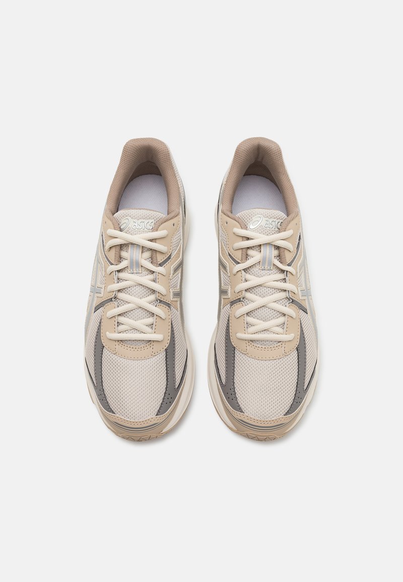 Zapatillas deportivas beige con parte superior de malla, acentos en gris claro y suelas texturizadas. Presentan un cierre con cordones en la parte delantera y un collar acolchado.