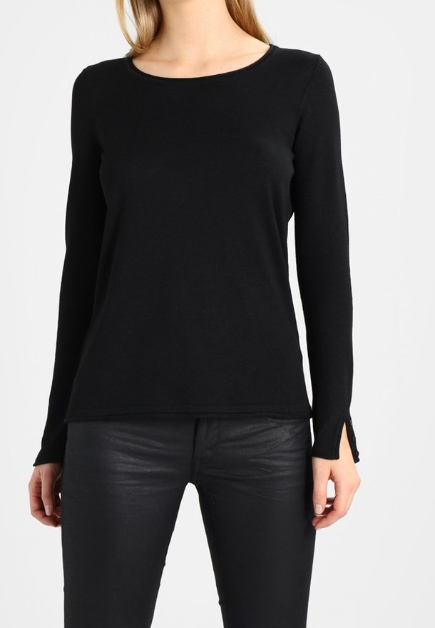 Femme portant un haut noir ajusté à manches longues avec de petits boutons aux poignets et un pantalon noir effet cuir, sur un fond clair uni.